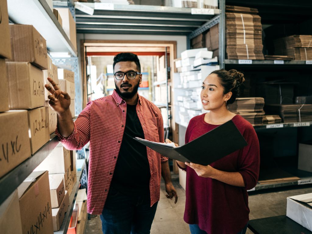 Succession Planning Isn’t Just For Big Companies- Here’s Why Your Small Business Needs One 3 Two individuals discuss a document while standing in a warehouse filled with neatly stacked boxes and storage racks.